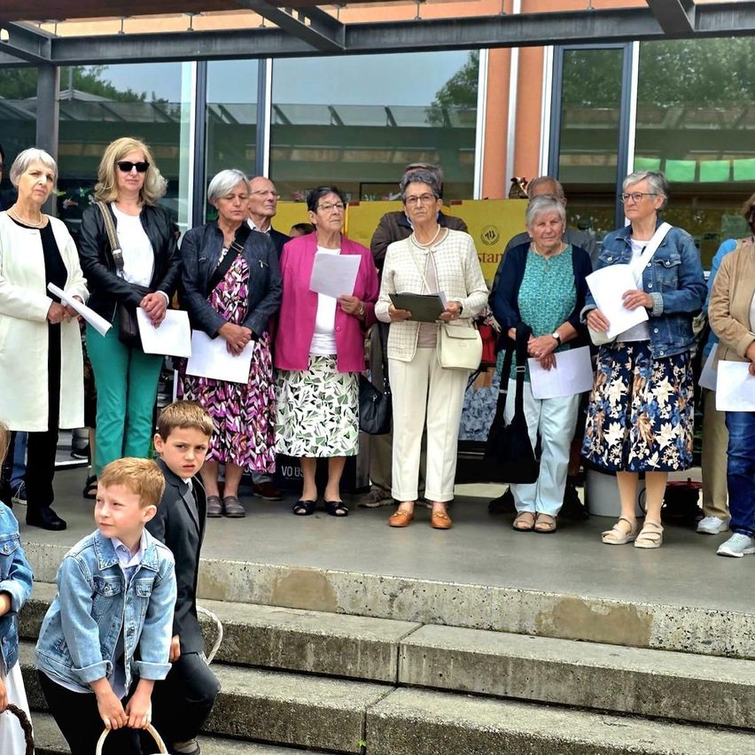 A group of elderly women and two boys are standing on a platform, holding papers and wearing glasses. The women are standing in a row and wearing various outfits. They appear to be attending a formal event.