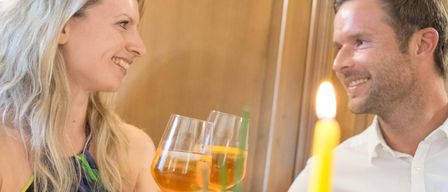 A couple, a man and a woman, smiling and holding wine glasses with a yellow liquid inside, in a dimly lit room. They are sitting next to each other, possibly in a restaurant.