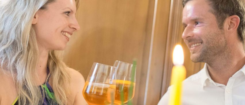 Ein Paar, ein Mann und eine Frau, lächeln und halten Weinglas mit gelber Flüssigkeit darin, in einem schwach beleuchteten Raum. Sie sitzen nebeneinander, wahrscheinlich in einem Restaurant.