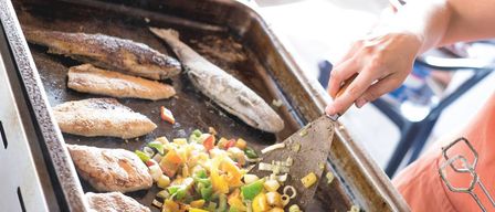 Eine Person benutzt einen Spachtel, um eine Pfanne mit Fisch und Gemüse zu rühren, einschließlich Paprika und Zwiebeln.