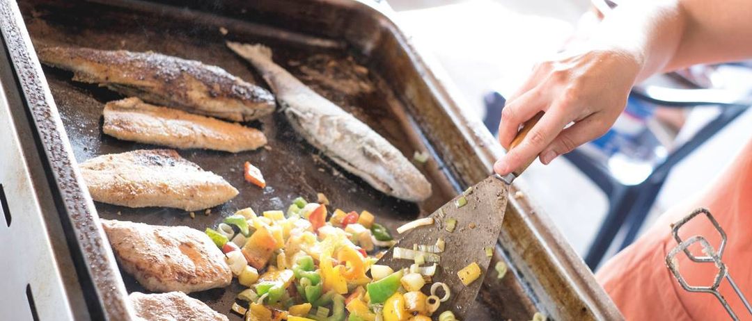 Eine Person benutzt einen Spachtel, um eine Pfanne mit Fisch und Gemüse zu rühren, einschließlich Paprika und Zwiebeln.