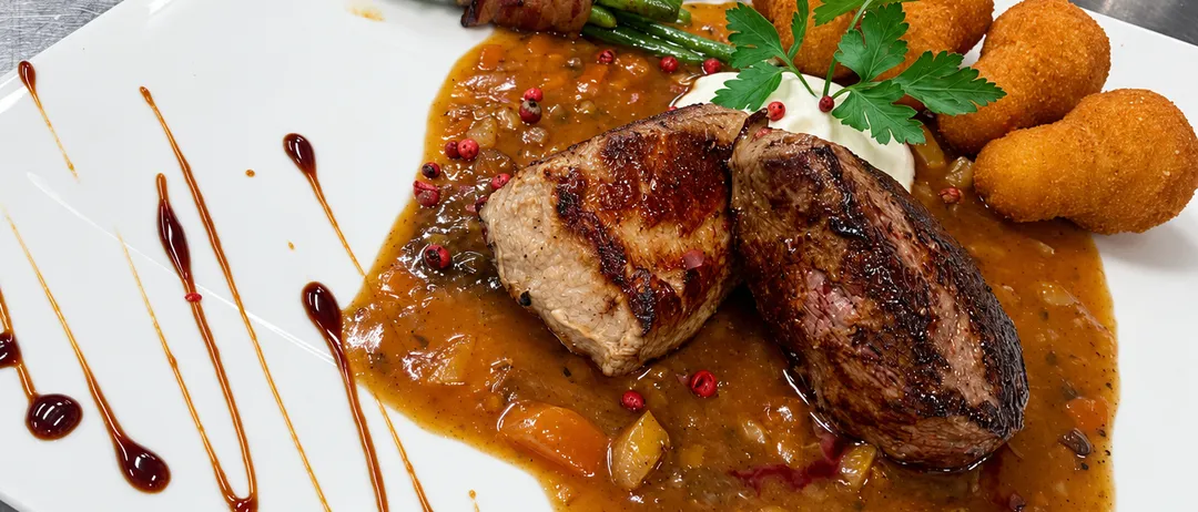 Ein Teller mit zwei Stücken Fleisch, einer Sauce, grünen Bohnen, die in Speck eingewickelt sind, und einigen frittierten Bällen.