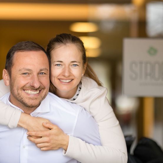 Ein lächelnder Mann und eine Frau umarmen sich in einem gemütlichen Innenraum. Die Frau steht hinter dem Mann, ihre Arme um seine Schultern. Sie sehen beide glücklich aus. Im Hintergrund steht ein Schild mit der Aufschrift 'STARK'.