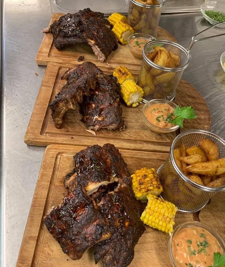 Vier Holzschneidebretter mit gegrillten Rippchen und Maiskolben. Ein Korb mit frittierten Kartoffeln und eine Schüssel Dip-Sauce sind in der Nähe. Die Umgebung ist eine Metalloberfläche.