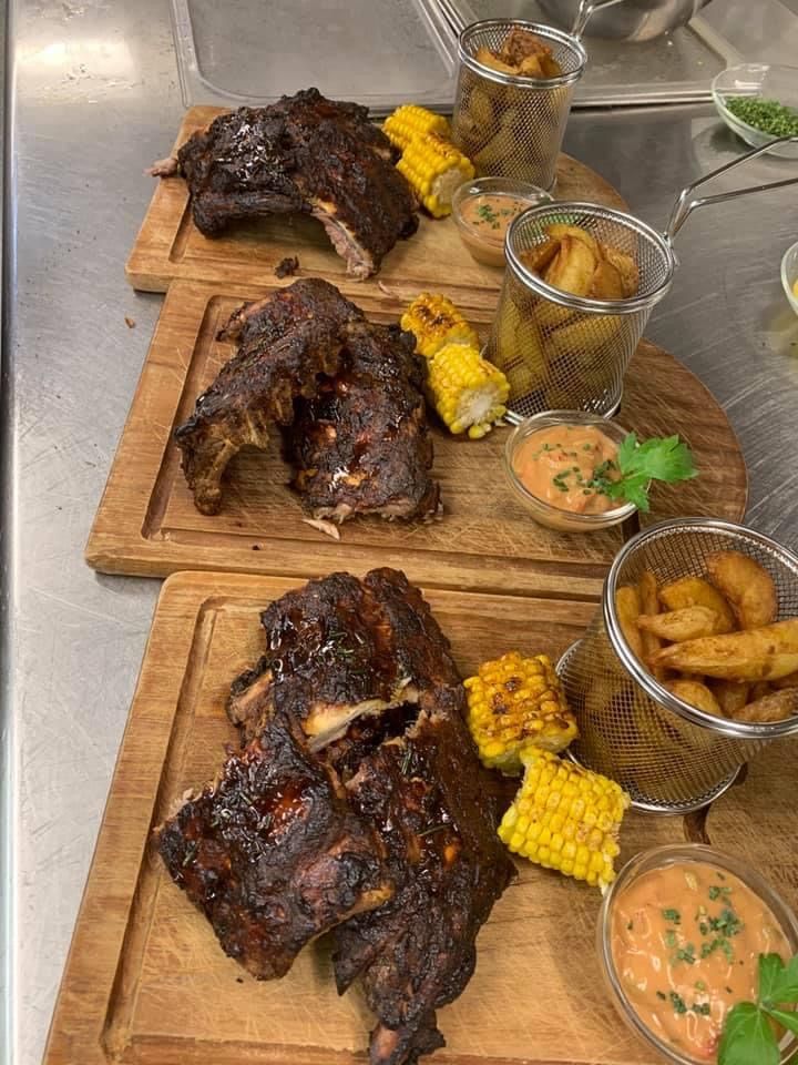 Three wooden cutting boards with grilled ribs, corn on the cob, and dipping sauces. The ribs are charred and juicy, garnished with herbs. The corn is golden and sweet. The sauces are in small bowls.