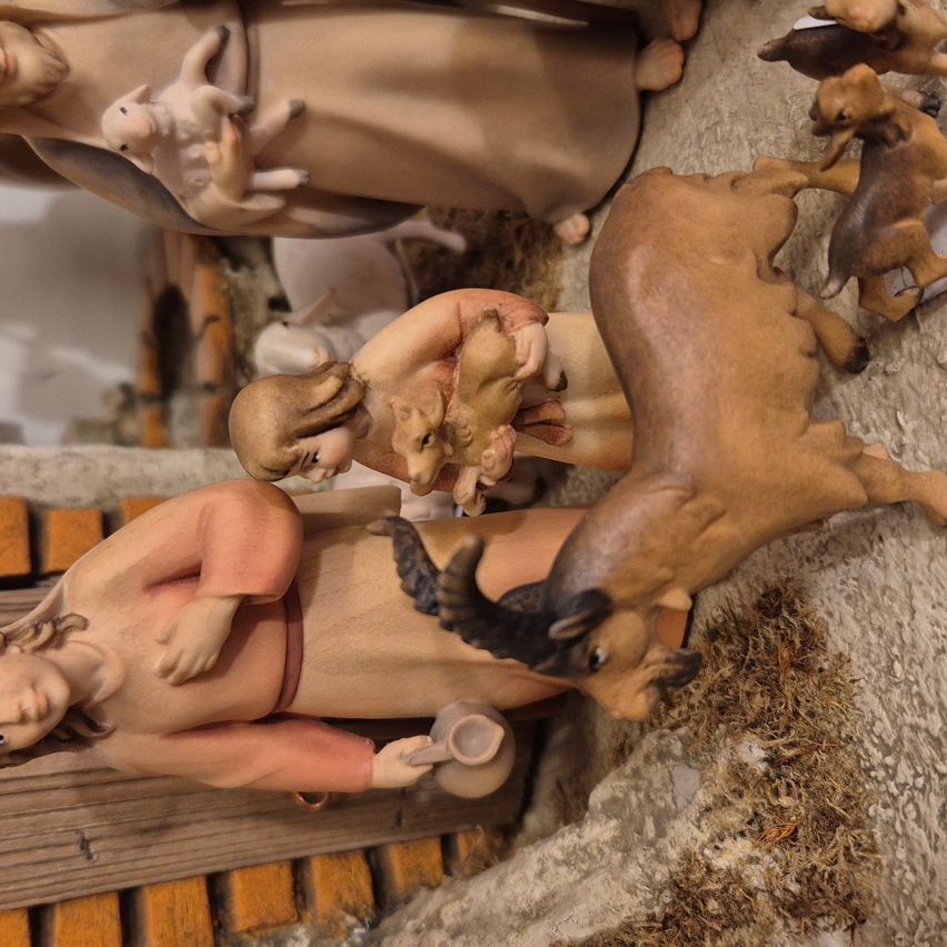 A nativity scene figurine of a woman feeding a baby lamb, surrounded by other figurines of animals and people, with a clay background.