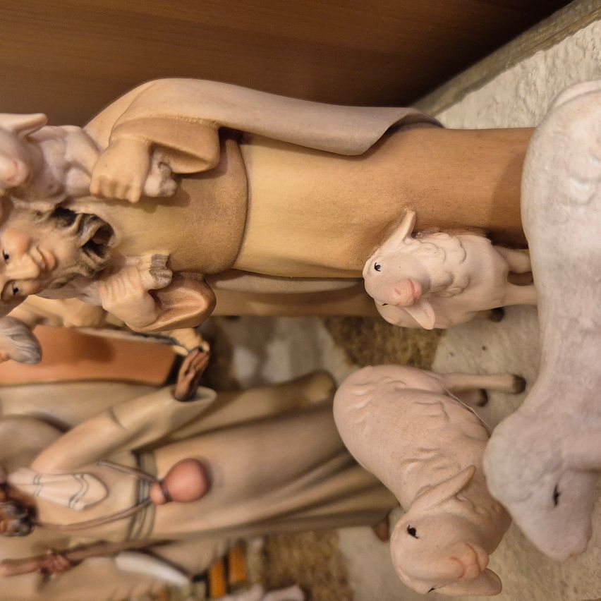 A close-up of a nativity scene with figures of Mary, Joseph, and baby Jesus. The baby Jesus is held by Joseph, while Mary holds another infant. Two lambs are placed beside them.