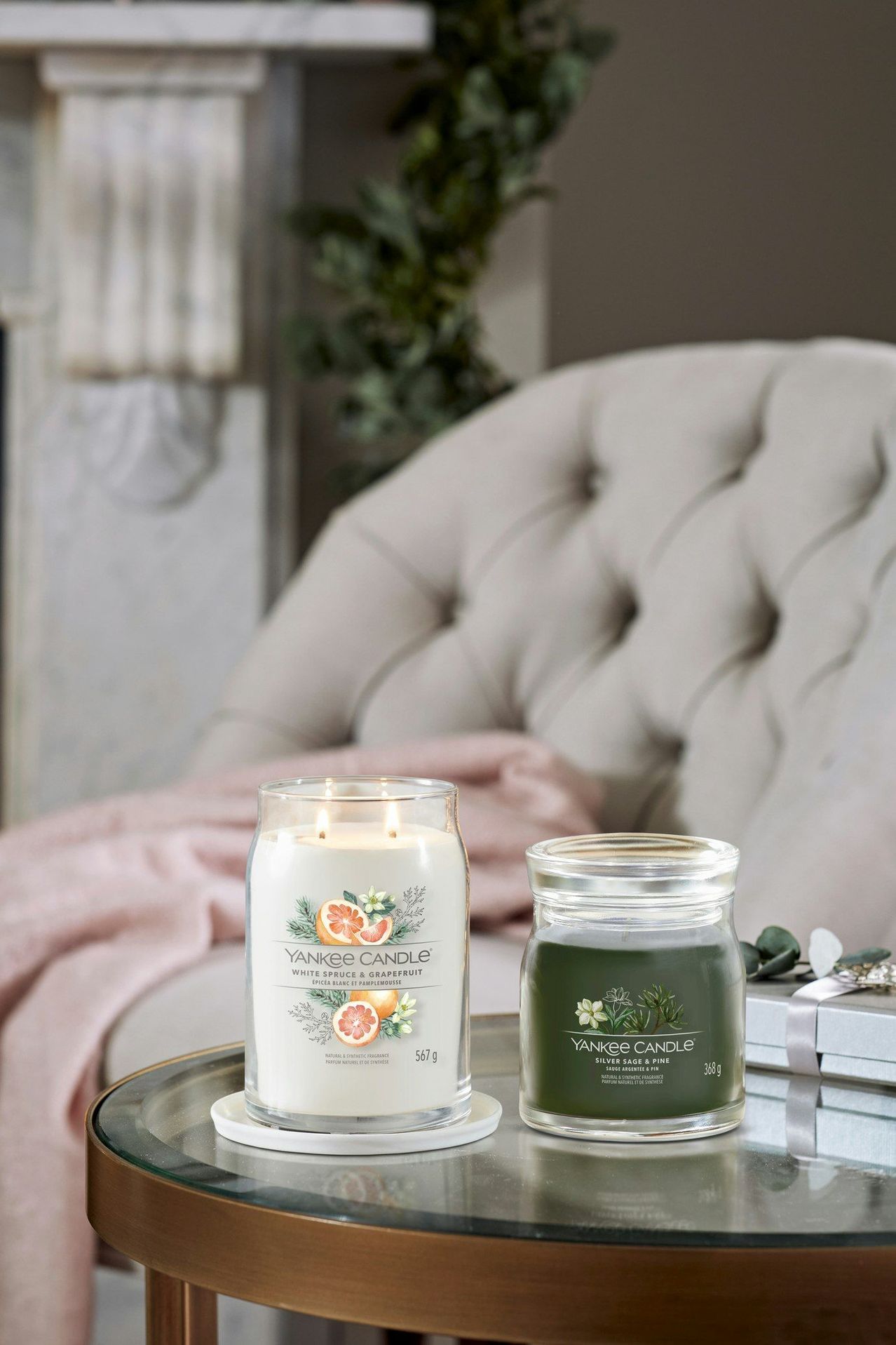 Two Yankee Candle jars are on a glass table. One is White Spruce & Grapefruit, and the other is Silver Sage & Pine. A chair with a pink blanket is in the background.