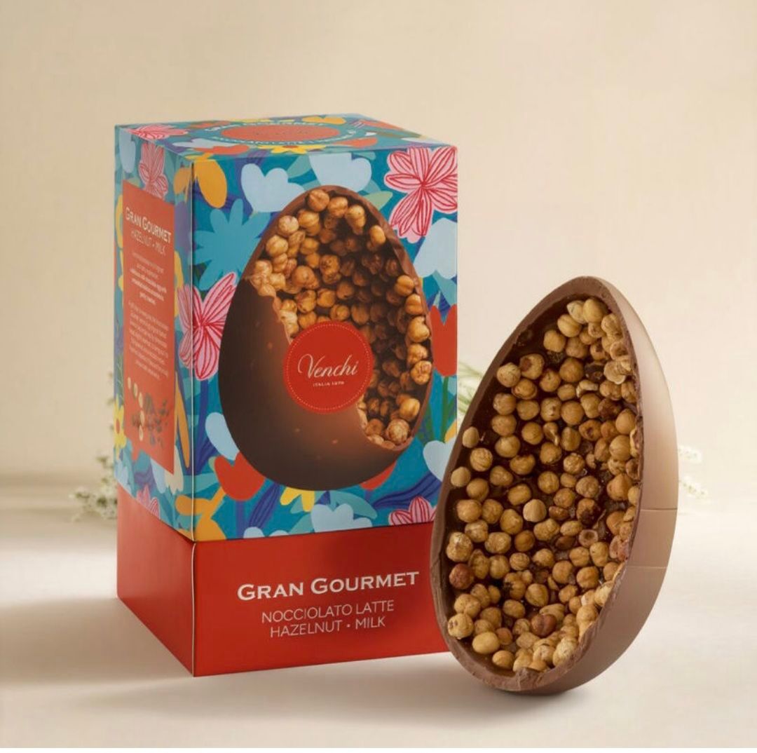 A large chocolate egg filled with hazelnuts, displayed next to its box, with colorful floral patterns. The box has the brand name 'Gran Gourmet' and the product name 'Nocciolato Latte'.