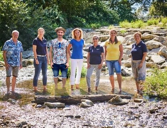 Sieben Menschen stehen in einem Bach, lächeln und posieren für ein Foto. Der Wasserstand ist niedrig, und sie sind von Felsen und Pflanzen umgeben.