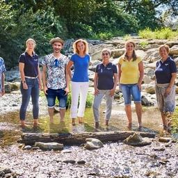 Sieben Menschen stehen in einem Bach, lächeln und posieren für ein Foto. Der Wasserstand ist niedrig, und sie sind von Felsen und Pflanzen umgeben.