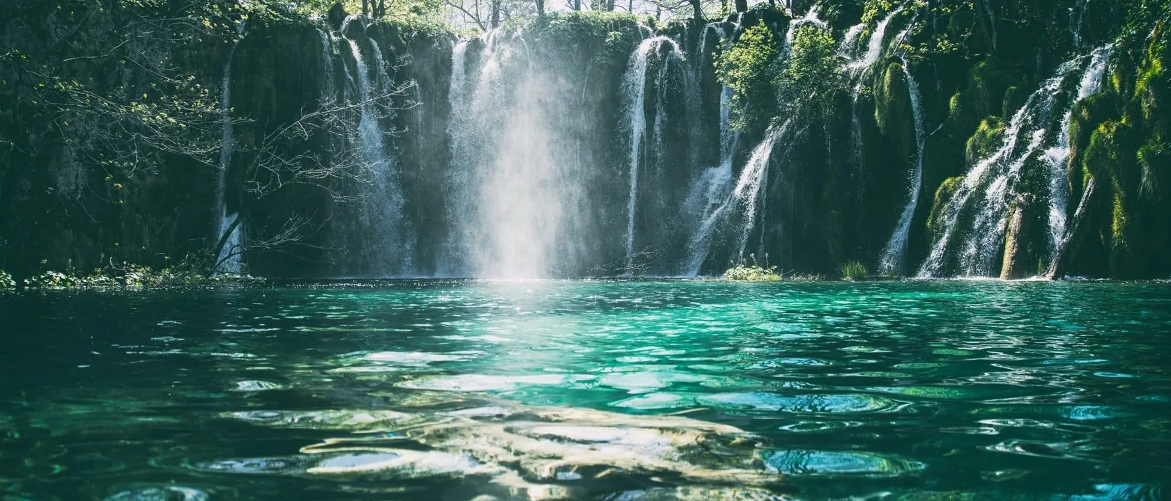 Ein atemberaubender Wasserfall mit kristallklarem türkisfarbenem Wasser, das über felsige Klippen in einen Pool fließt, umgeben von üppigem Grün und Bäumen.