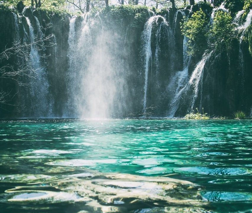 Ein atemberaubender Wasserfall mit kristallklarem türkisfarbenem Wasser, das über felsige Klippen in einen Pool fließt, umgeben von üppigem Grün und Bäumen.
