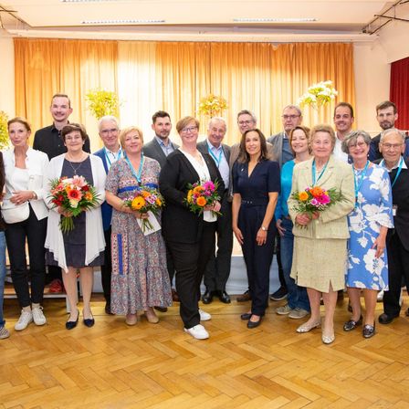 Eine Gruppe von Menschen in formeller Kleidung steht zusammen und hält Blumen, wahrscheinlich bei einer Veranstaltung. Sie sind vor einer Kulisse mit gelben Vorhängen und einem Tisch mit Blumen angeordnet.