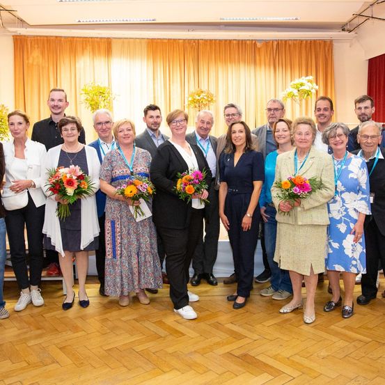 Eine Gruppe von Menschen in formeller Kleidung steht zusammen und hält Blumen, wahrscheinlich bei einer Veranstaltung. Sie sind vor einer Kulisse mit gelben Vorhängen und einem Tisch mit Blumen angeordnet.