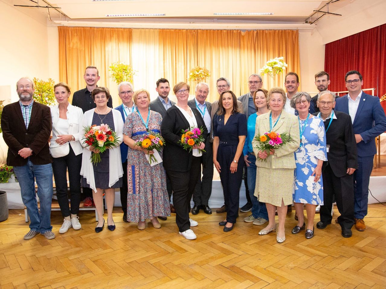 Eine Gruppe von Menschen in formeller Kleidung steht zusammen und hält Blumen, wahrscheinlich bei einer Veranstaltung. Sie sind vor einer Kulisse mit gelben Vorhängen und einem Tisch mit Blumen angeordnet.