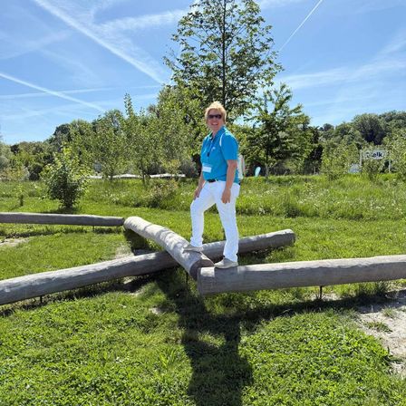 Eine Frau in einem blauen Shirt und weißen Hosen steht auf einem Holzstamm in einem Grasfeld. Bäume und Pflanzen umgeben den Bereich.