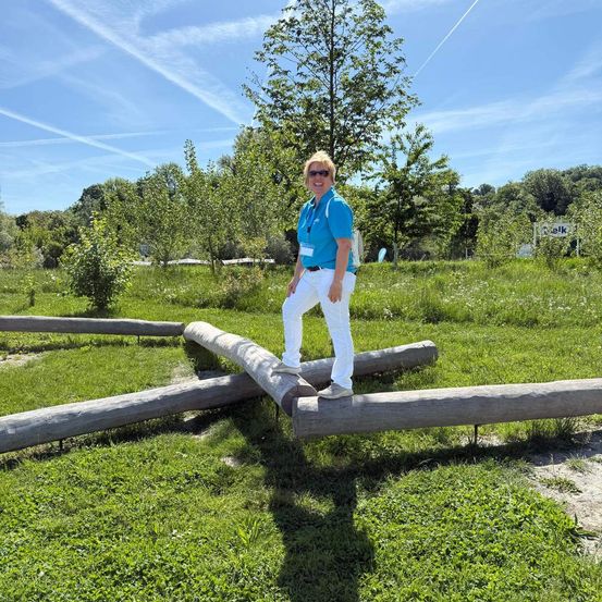 Eine Frau in einem blauen Shirt und weißen Hosen steht auf einem Holzstamm in einem Grasfeld. Bäume und Pflanzen umgeben den Bereich.