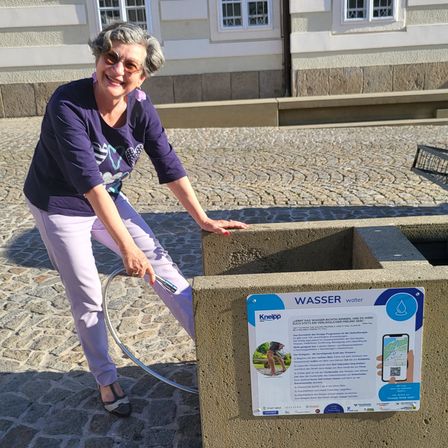 Eine lächelnde Frau in lila Kleidung füllt eine Wasserflasche an einem Betonbrunnen auf. Hinter ihr steht ein Gebäude mit weißen Fenstern und einem Schild, das 'Wasser' steht.