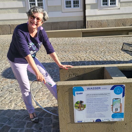 Eine lächelnde Frau in lila Kleidung füllt eine Wasserflasche an einem Betonbrunnen auf. Hinter ihr steht ein Gebäude mit weißen Fenstern und einem Schild, das 'Wasser' steht.