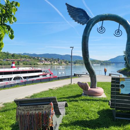 Ein ruhiger Parkblick mit einer großen Skulptur, die einem Fisch mit Flügeln ähnelt. Bänke sind unter einem Baum platziert, und eine Person steht am Ufer. Ein Boot ist im Fluss verankert, und Berge sind in der Ferne sichtbar.