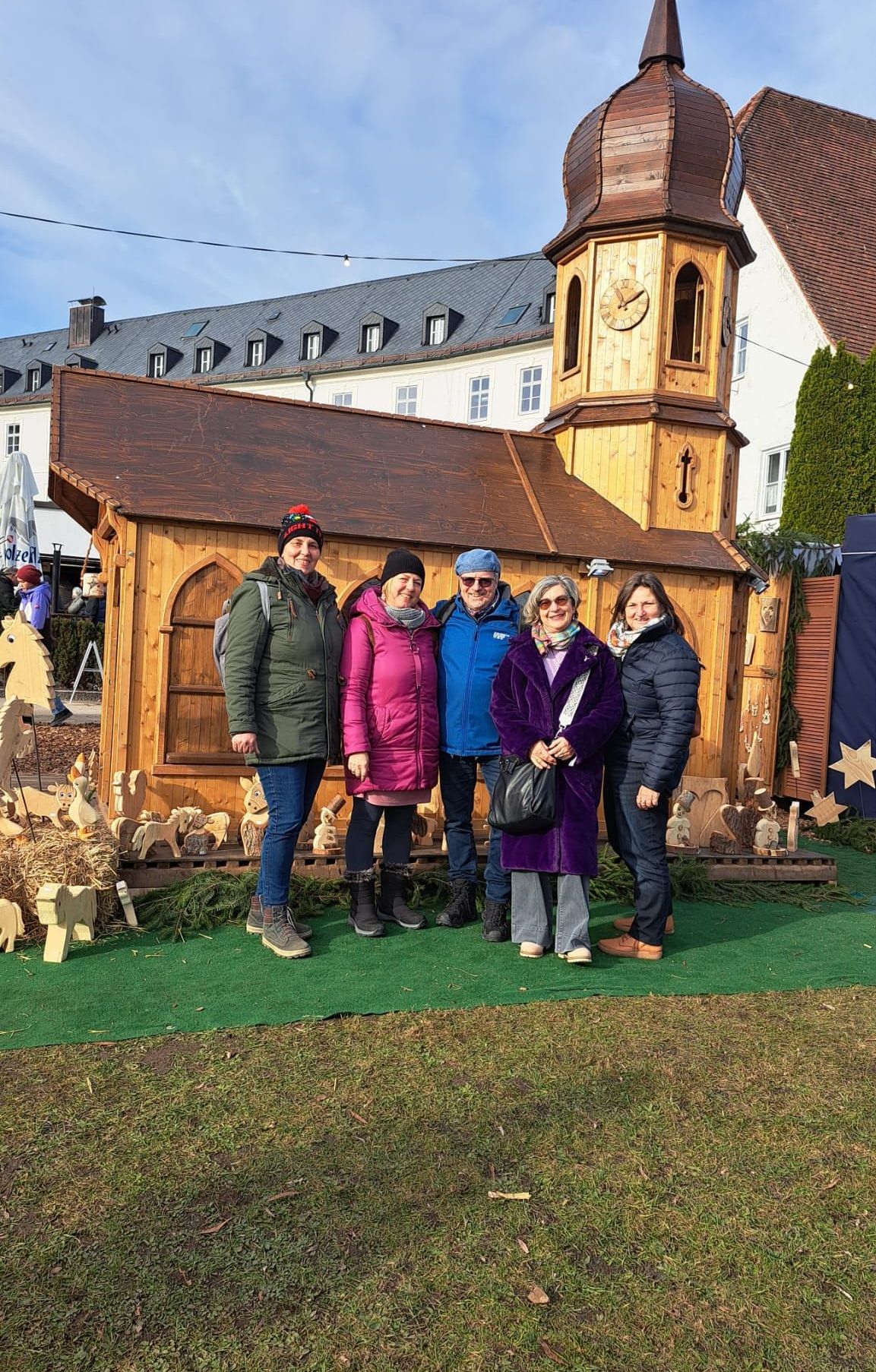 Eine Gruppe von Menschen in Winterkleidung posiert vor einem hölzernen Kirchenmodell mit einem Glockenturm. Sie stehen auf grünem Gras mit geschnitzten Holztieren in der Nähe.