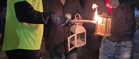 Drei Männer halten brennende Laternen in der Hand und stehen nachts im Freien, mit Menschen und Gebäuden im Hintergrund.