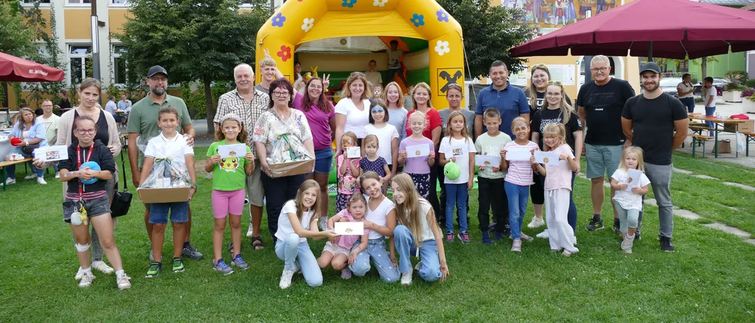 Eine Gruppe von Erwachsenen und Kindern posiert für ein Foto vor einer aufblasbaren Burg, einige halten Auszeichnungen und andere stehen in der Nähe von Bäumen und einer Wandmalerei.