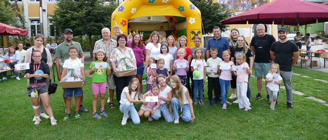 Eine Gruppe von Erwachsenen und Kindern posiert für ein Foto vor einer aufblasbaren Burg, einige halten Auszeichnungen und andere stehen in der Nähe von Bäumen und einer Wandmalerei.