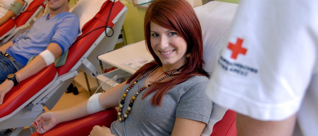 Eine Frau mit roten Haaren lächelt, während sie auf einem roten Stuhl in einem Blutspendezentrum sitzt. Ein Mann sitzt hinter ihr auf einem Stuhl.