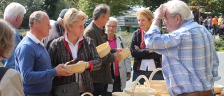 Mehrere Personen stehen um einen Tisch im Freien und betrachten gewebte Körbe. Eine Frau hält einen Korb, während ein anderer Mann auf einen zeigt. Im Hintergrund befinden sich Bäume und ein Gebäude.