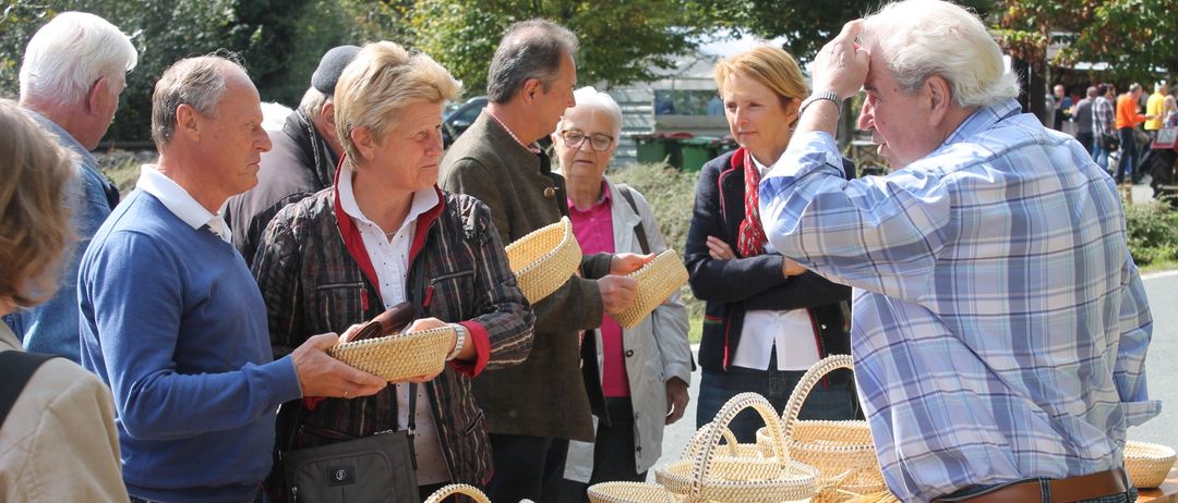 Mehrere Personen stehen um einen Tisch im Freien und betrachten gewebte Körbe. Eine Frau hält einen Korb, während ein anderer Mann auf einen zeigt. Im Hintergrund befinden sich Bäume und ein Gebäude.
