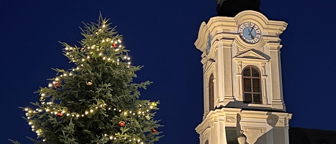 Ein großer Weihnachtsbaum mit bunten Ornamenten steht vor einem hohen weißen Glockenturm. Der Baum ist mit weißen und roten Lichtern beleuchtet. Der Turm hat zwei Uhren, eine oben und eine an der Seite. Der Himmel ist dunkel.