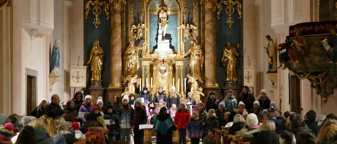 Ein Chor von Kindern in warmen Winterkleidung singt in einer Kirche, mit einem kunstvollen Altar im Hintergrund und Menschen, die auf hölzernen Kirchenbänken sitzen.