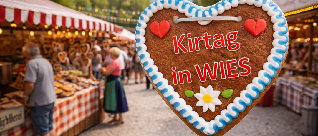 Ein herzförmiger Keks mit blauer und weißer Glasur und rotem Text 'Kirtaag in WIES' hängt vor einem geschäftigen Markt mit Ständen und Menschen.