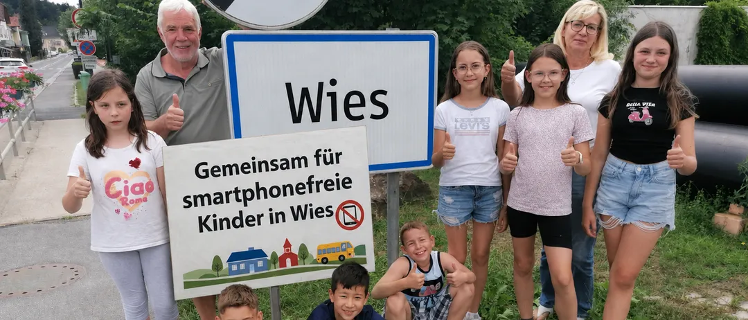 Eine Gruppe von Kindern und Erwachsenen posiert für ein Foto vor einem Schild mit der Aufschrift 'Wies' und einem Banner, das 'Gemeinsam für smartphonefreie Kinder in Wies' sagt. Sie alle zeigen Daumen hoch.