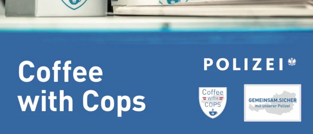 Zwei Polizeibeamte stehen hinter einem Tisch mit Kaffeetassen und Schachteln, die mit 'Kaffee mit Cops' beschriftet sind.