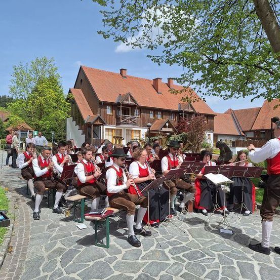 Ein Orchester spielt im Freien in traditioneller deutscher Kleidung vor Häusern mit roten Dächern.