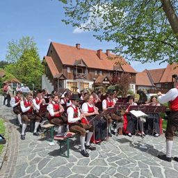Ein Orchester spielt im Freien in traditioneller deutscher Kleidung vor Häusern mit roten Dächern.