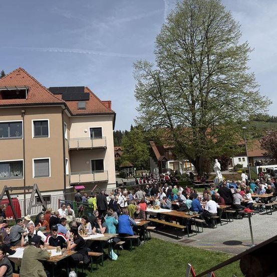 Eine große Menschenmenge versammelt sich auf einer Außensitzfläche und genießt Essen und Getränke an Picknicktischen unter einem klaren Himmel. Die Umgebung umfasst ein Gebäude mit Solarmodulen und einen großen Baum im Hintergrund.