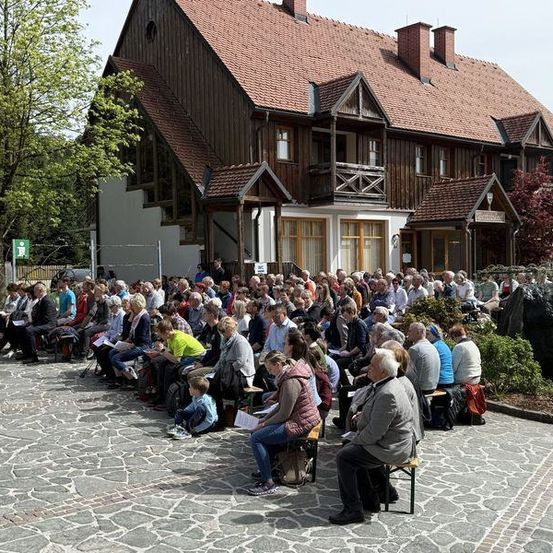 Eine große Menschenmenge sitzt auf Steinbänken vor einem traditionellen Holzhaus. Einige halten Papiere, andere tragen Turnschuhe. Ein Baum und ein Schild befinden sich auf der linken Seite.