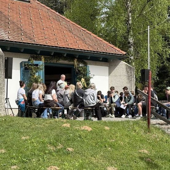 Eine Gruppe von Menschen sitzt auf Bänken vor einem Gebäude mit rotem Dach. Ein Mann steht vor ihnen und spricht. Im Hintergrund befinden sich Bäume und eine Fahne.