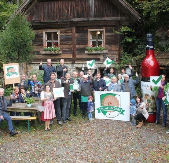 Eine Gruppe von Menschen steht vor einem rustikalen Haus, einige mit Schildern, und eine große Flasche im Hintergrund. Die Menschen lächeln und posieren für ein Foto.