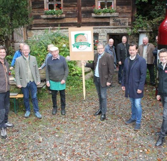 Eine Gruppe von Männern steht vor einem rustikalen Gebäude mit einem Holzschild und lächelt für die Kamera. Dahinter ist eine große Flasche auffällig angebracht.
