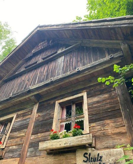 Ein altes Holzhaus mit einem geneigten Dach und einem Schild mit der Aufschrift 'Stutz Mühle'. Es gibt zwei Fenster mit roten Blumen auf der Fensterbank.