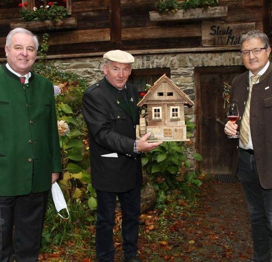 Drei Männer stehen vor einem Holzhaus. Der Mann in der Mitte hält ein Miniaturhaus und der Mann auf der rechten Seite hält ein Weinglas.