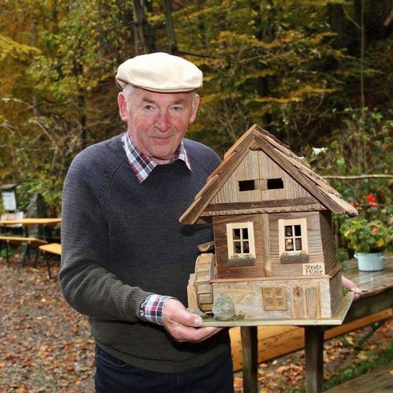 Ein älterer Mann hält ein kleines Holzhaus mit einem Rad vor einem herbstlichen Waldhintergrund. Er trägt eine Baskenmütze und einen grauen Pullover.