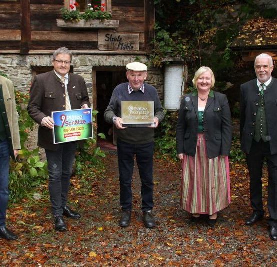 Fünf Personen stehen vor einem Gebäude, lächeln und posieren für ein Foto. Zwei Männer halten Trophäen und eine Frau hält ein Plakat. Hinter ihnen sind Pflanzen und ein Gebäude.