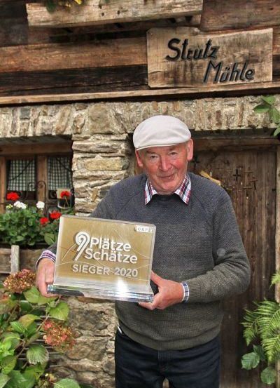 Ein Mann in einem Hut steht vor einem Steinhaus namens Stutz Muhle. Er hält eine Trophäe mit der Aufschrift 9 Platze Schatze Sieger 2020.