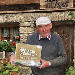 Ein Mann in einem Hut steht vor einem Steinhaus namens Stutz Muhle. Er hält eine Trophäe mit der Aufschrift 9 Platze Schatze Sieger 2020.